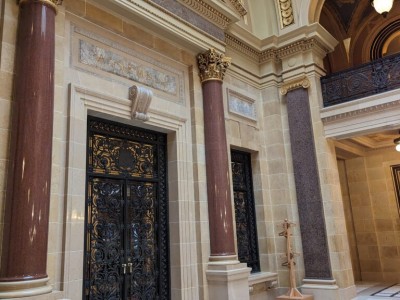 An ornate stone doorway with dark, intricate iron gates flanked by polished brown marble columns beneath a marble "ASSEMBLY" sign.