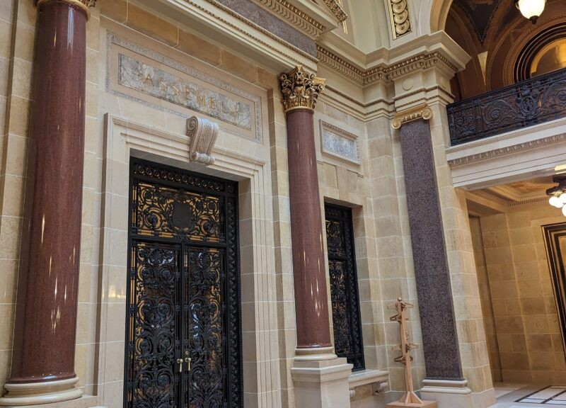 An ornate stone doorway with dark, intricate iron gates flanked by polished brown marble columns beneath a marble "ASSEMBLY" sign.