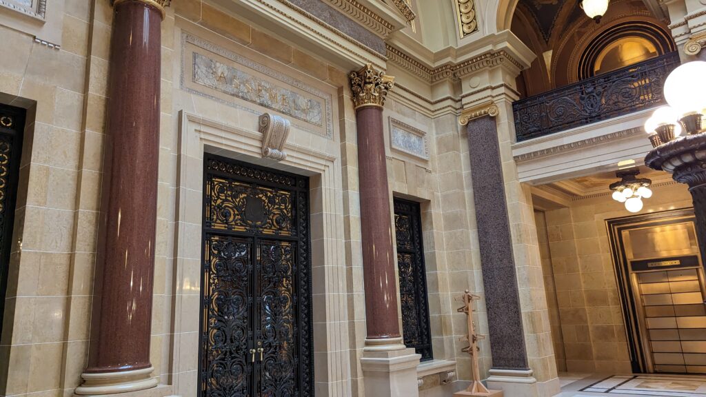 An ornate stone doorway with dark, intricate iron gates flanked by polished brown marble columns beneath a marble "ASSEMBLY" sign.