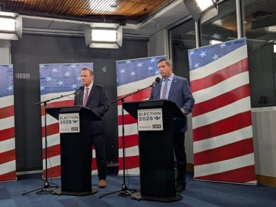 Two men in suits stand at separate "ELECTION 2026" podiums in front of American flags during a televised debate.