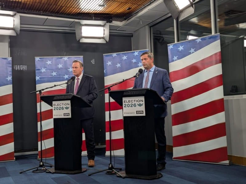 Two men in suits stand at separate "ELECTION 2026" podiums in front of American flags during a televised debate.