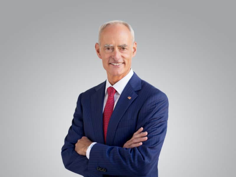 Ron Schutz poses with his arms crossed in a blue pinstripe suit, red patterned tie, and small U.S. flag lapel pin.