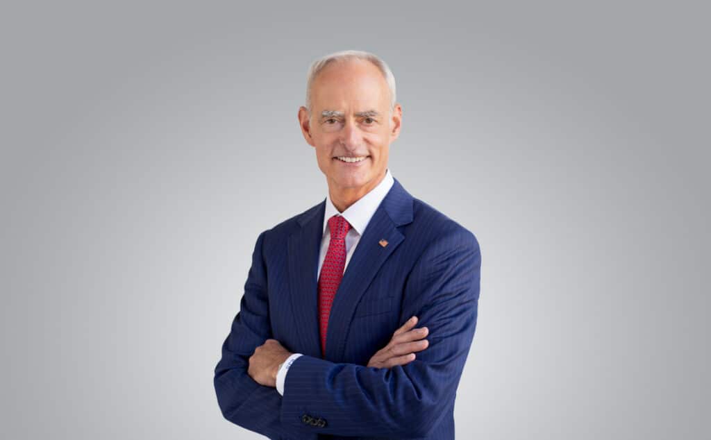 Ron Schutz poses with his arms crossed in a blue pinstripe suit, red patterned tie, and small U.S. flag lapel pin.