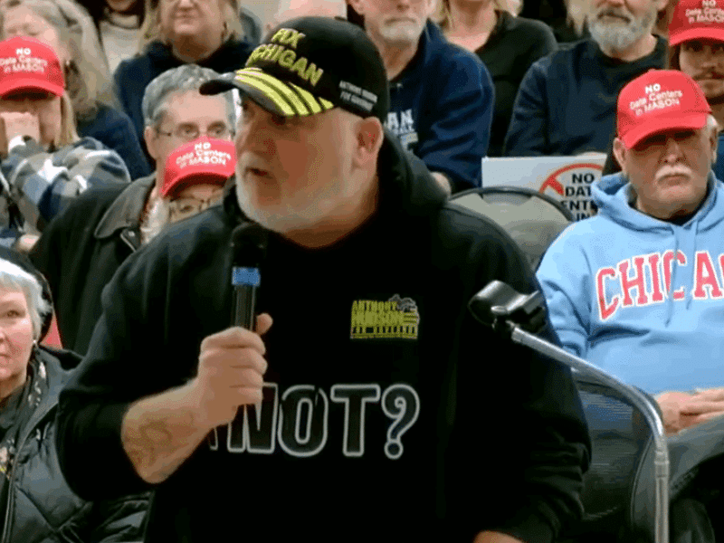 Michigan gubernatorial candidate Anthony Hudson speaks into a microphone at a city council meeting, wearing a black sweatshirt reading "[illegible] NOT?" and a cap reading "FIX MICHIGAN."