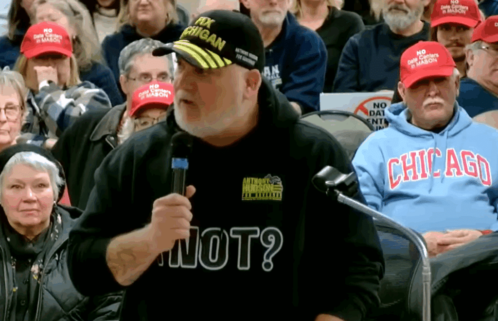 Michigan gubernatorial candidate Anthony Hudson speaks into a microphone at a city council meeting, wearing a black sweatshirt reading "[illegible] NOT?" and a cap reading "FIX MICHIGAN."
