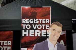 A sign reading "REGISTER TO VOTE HERE!" stands next to a smiling cardboard cutout of Charlie Kirk under a black canopy tent.