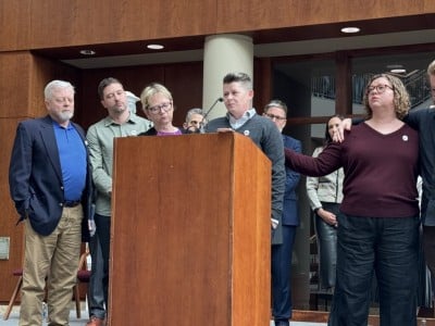 Becca Good stands at a wooden podium speaking into a microphone, surrounded by supportive family members and supporters.