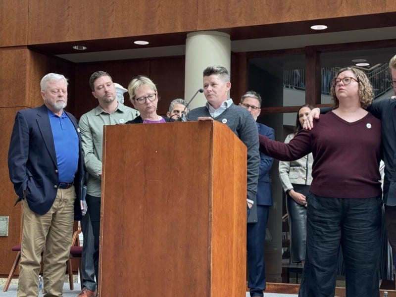 Becca Good stands at a wooden podium speaking into a microphone, surrounded by supportive family members and supporters.