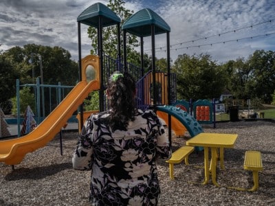 A woman with her back to the camera stands facing an empty outdoor playground with slides and a yellow picnic table.