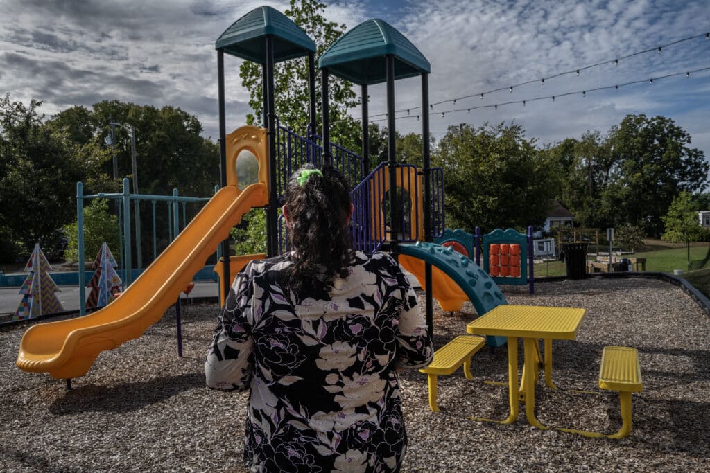A woman with her back to the camera stands facing an empty outdoor playground with slides and a yellow picnic table.