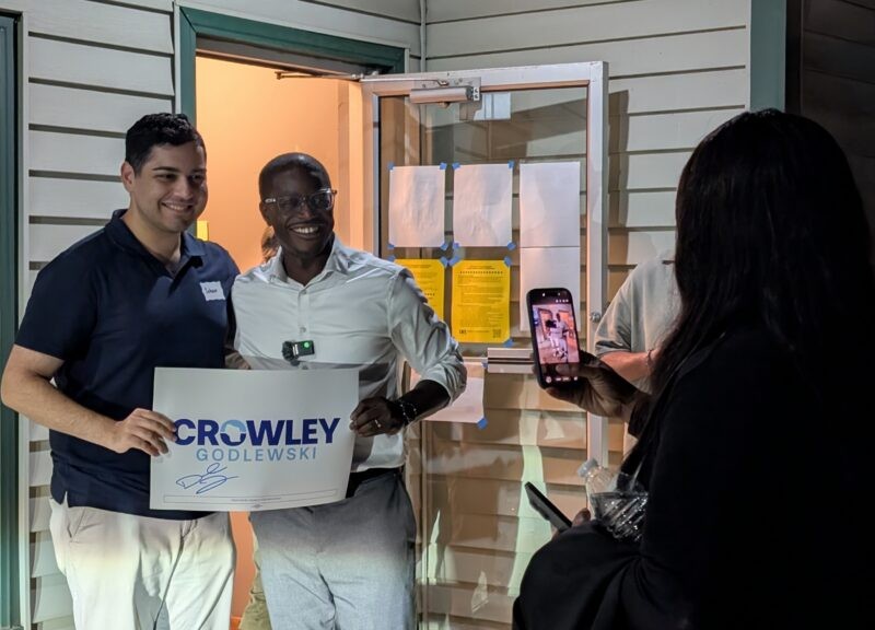 David Crowley and Soham Bhatnagar pose together holding a campaign sign as someone takes their picture with a smartphone.