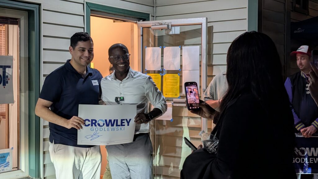 David Crowley and Soham Bhatnagar pose together holding a campaign sign as someone takes their picture with a smartphone.