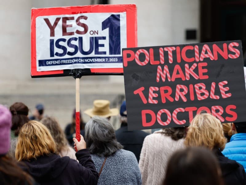 Demonstrators hold signs reading "YES ON ISSUE 1" and "POLITICIANS MAKE TERRIBLE DOCTORS" at an outdoor rally.