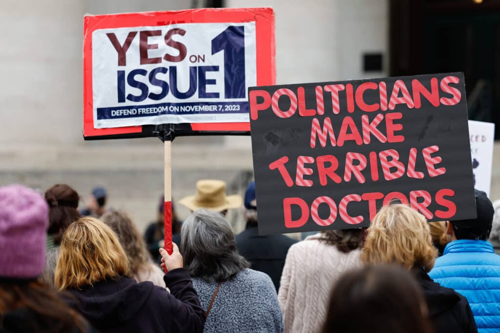 Demonstrators hold signs reading "YES ON ISSUE 1" and "POLITICIANS MAKE TERRIBLE DOCTORS" at an outdoor rally.