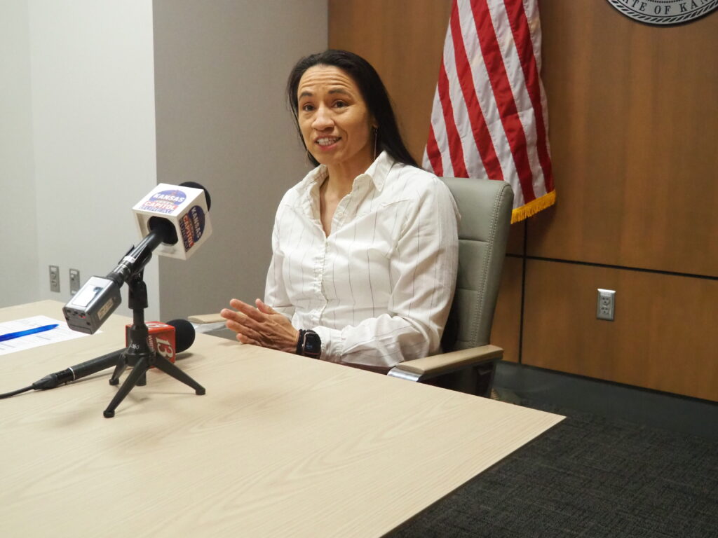 U.S. Rep. Sharice Davids speaks at a press conference table behind multiple microphones and in front of an American flag.