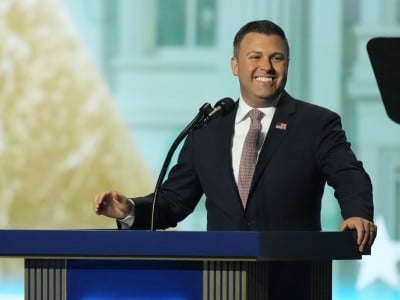 Rep. Ryan Mackenzie stands at a blue podium with a microphone, smiling while speaking.