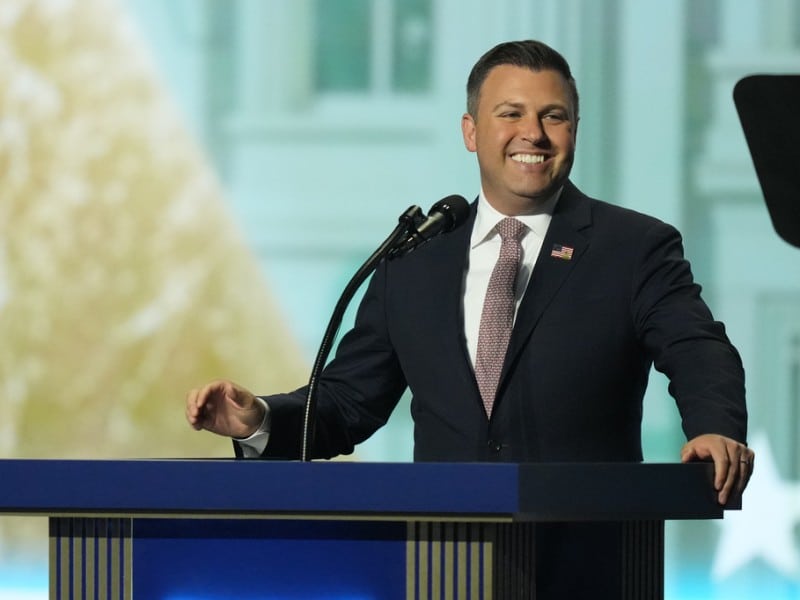 Rep. Ryan Mackenzie stands at a blue podium with a microphone, smiling while speaking.