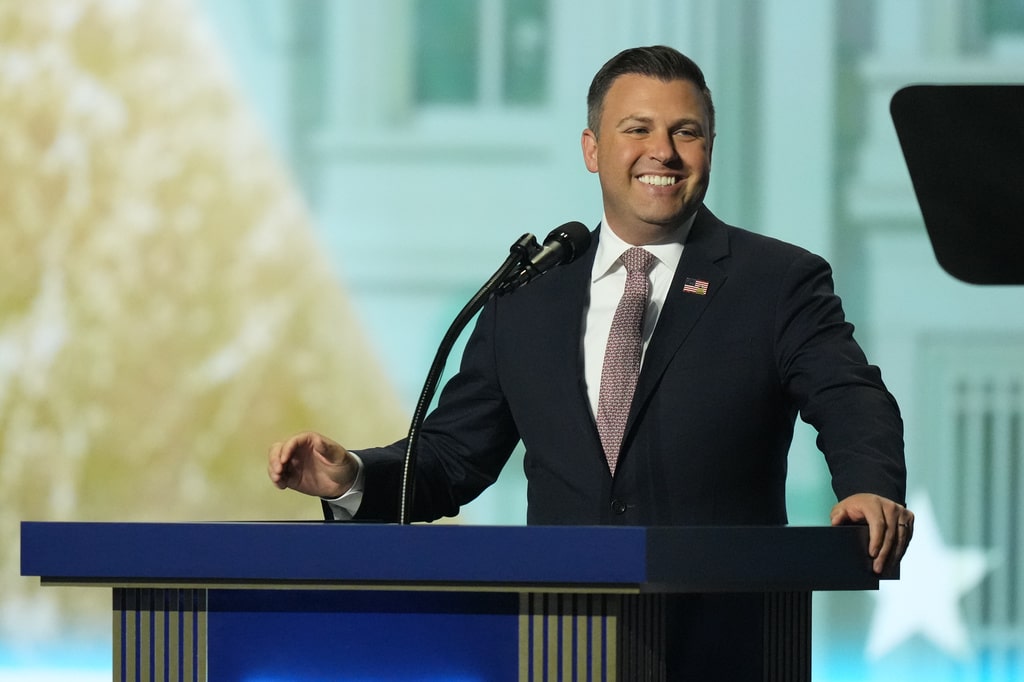 Rep. Ryan Mackenzie stands at a blue podium with a microphone, smiling while speaking.