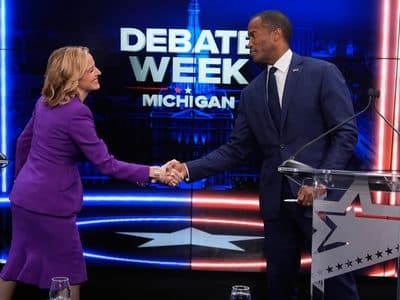 Jocelyn Benson and John James smile as they shake hands on a debate stage in front of a "DEBATE WEEK MICHIGAN" backdrop.