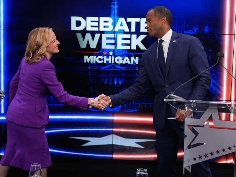 Jocelyn Benson and John James smile as they shake hands on a debate stage in front of a "DEBATE WEEK MICHIGAN" backdrop.