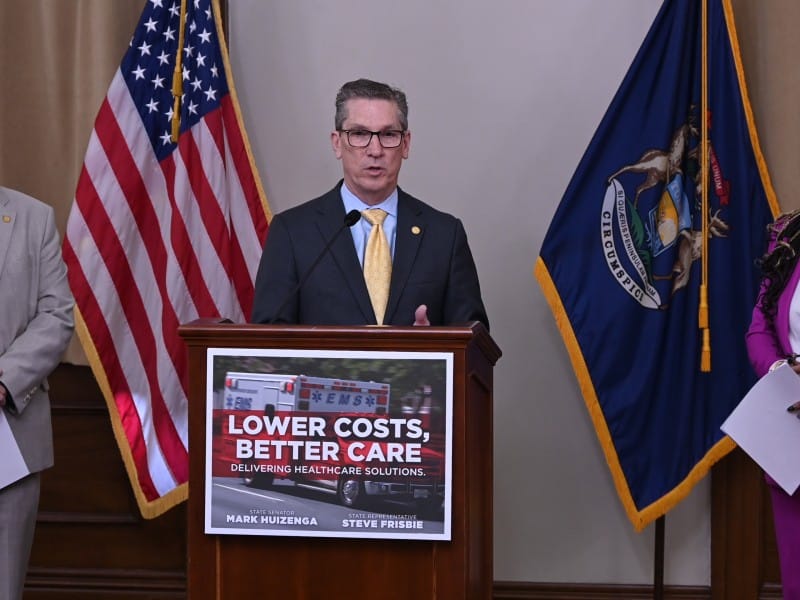 A man speaks at podium reading "Lower Costs, Better Care, Delivering Healthcare Solutions," flanked by colleagues and flags