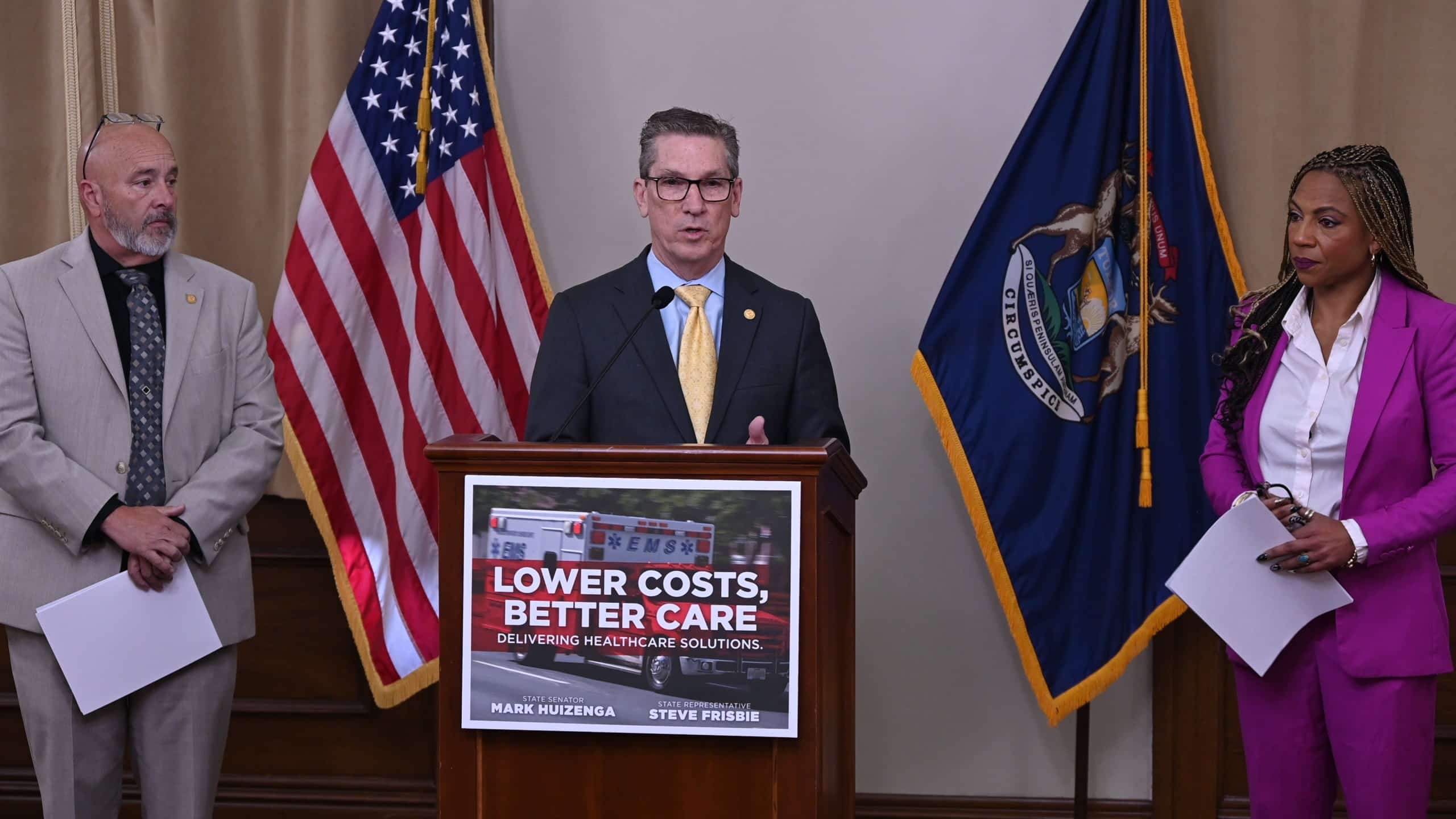 A man speaks at podium reading "Lower Costs, Better Care, Delivering Healthcare Solutions," flanked by colleagues and flags