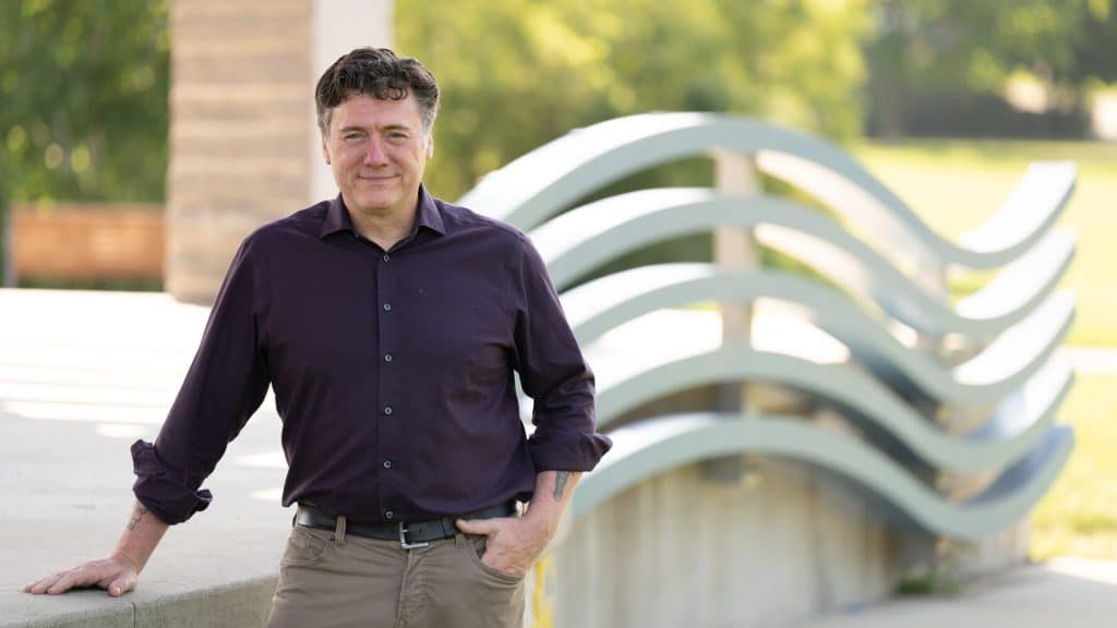 Aric Putnam in a purple shirt smiling outdoors, leaning against a ledge near a modern arched structure