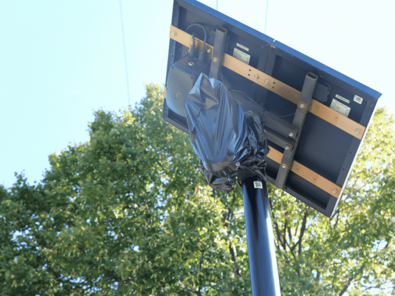 A solar panel and surveillance camera assembly mounted on a high pole, with the camera head wrapped in a black plastic bag.