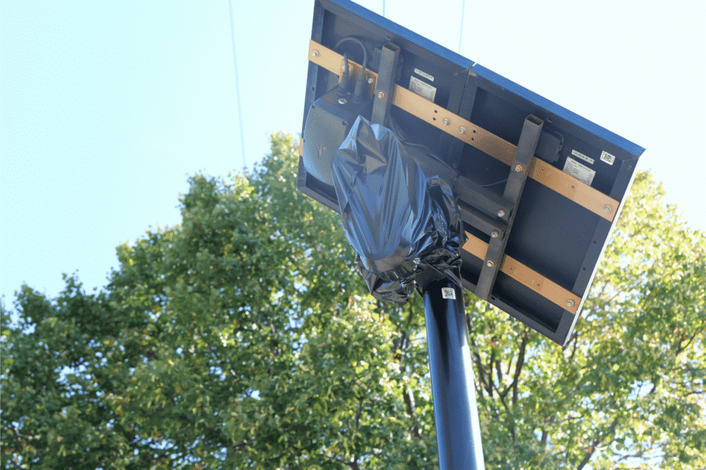 A solar panel and surveillance camera assembly mounted on a high pole, with the camera head wrapped in a black plastic bag.