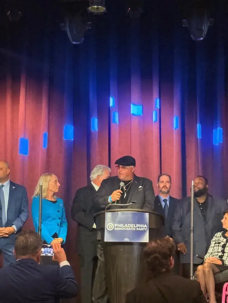 A man wearing a black flat cap and sunglasses speaks at a Philadelphia Democratic Party podium, flanked by other people on stage.