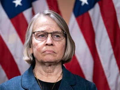 Woman with gray bob haircut and glasses stands solemnly in front of American flags