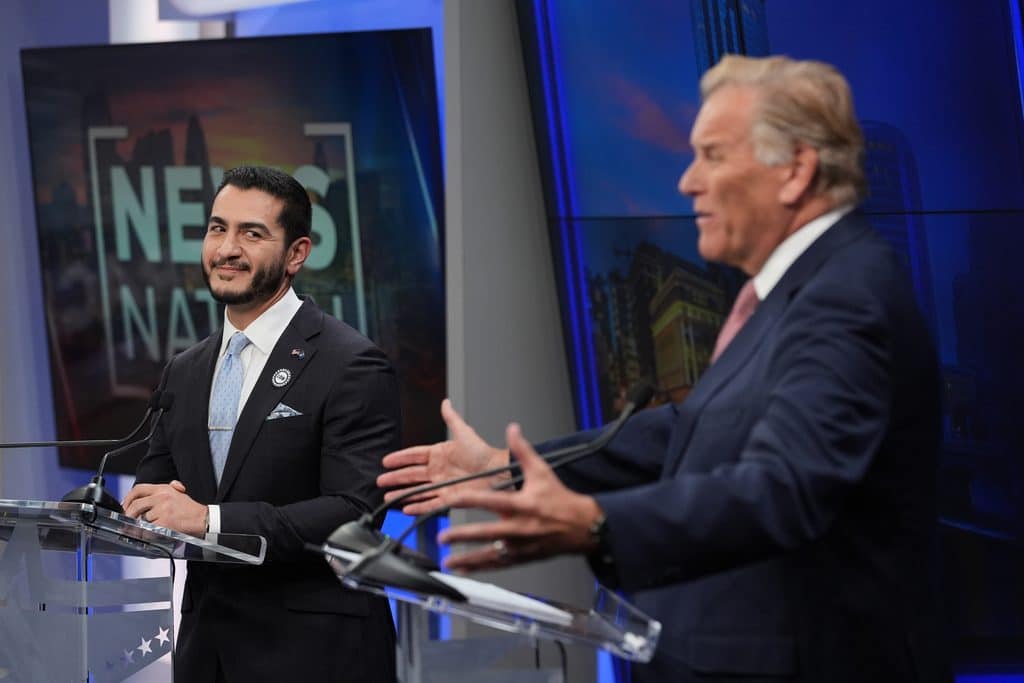 Abdul El-Sayed and Mike Rogers, both candidates for Michigan's open U.S. Senate seat, stand before clear podiums in a news studio. El-Sayed is on the left and smiling, Rogers is on the right, slightly out of focus, and speaking. 