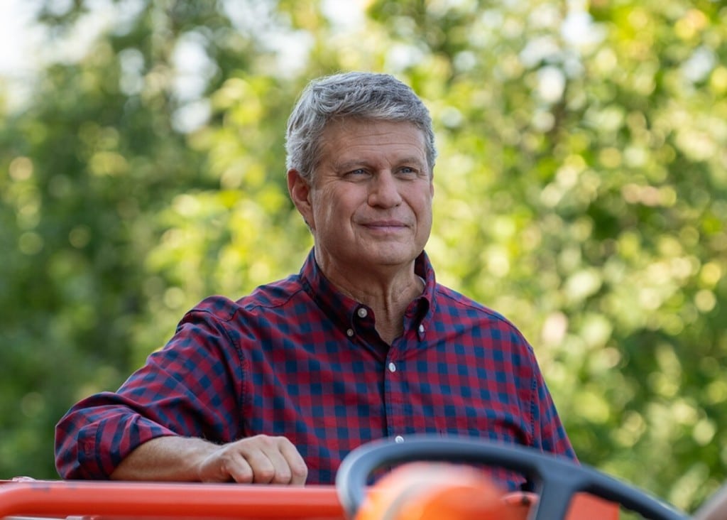 U.S. Rep. Bill Huizenga in a red and navy checked shirt sitting on an orange tractor outdoors, smiling