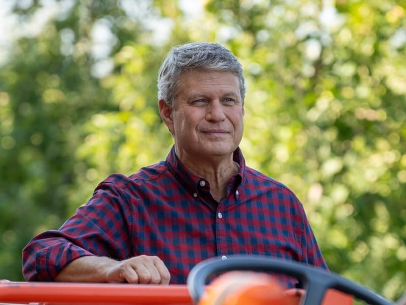 U.S. Rep. Bill Huizenga in a red and navy checked shirt sitting on an orange tractor outdoors, smiling