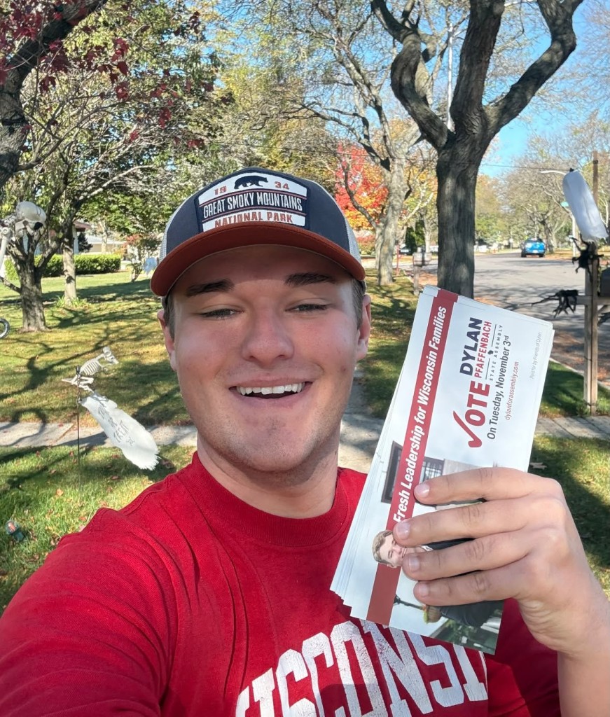 Dylan Pfaffenbach smiles outdoors while holding up a stack of campaign flyers on a sunny residential street.