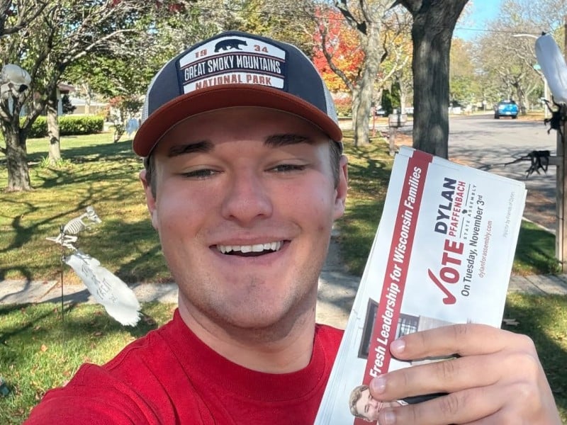 Dylan Pfaffenbach smiles outdoors while holding up a stack of campaign flyers on a sunny residential street.