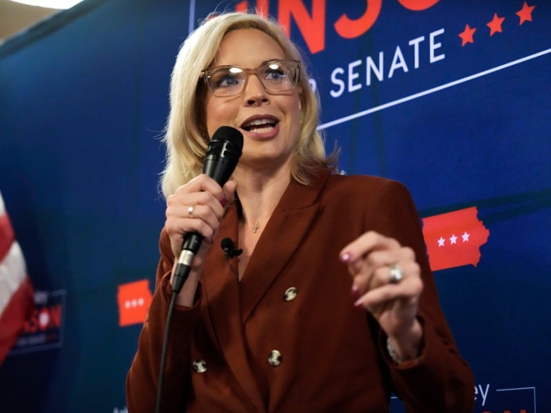 Rep. Ashley Hinson speaks into a handheld microphone in front of a blue campaign backdrop.