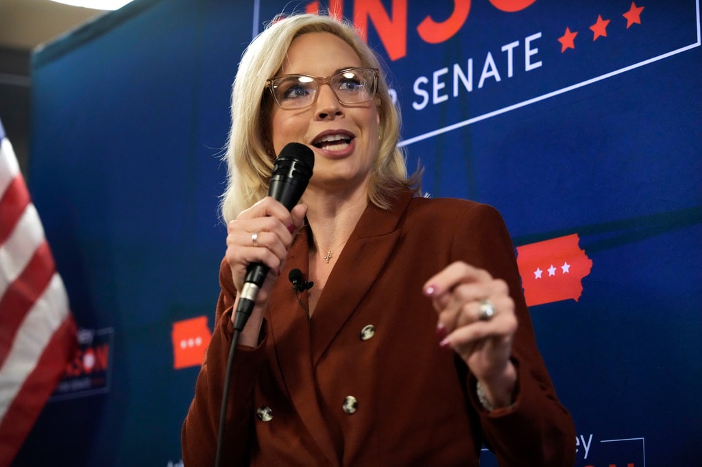 Rep. Ashley Hinson speaks into a handheld microphone in front of a blue campaign backdrop.
