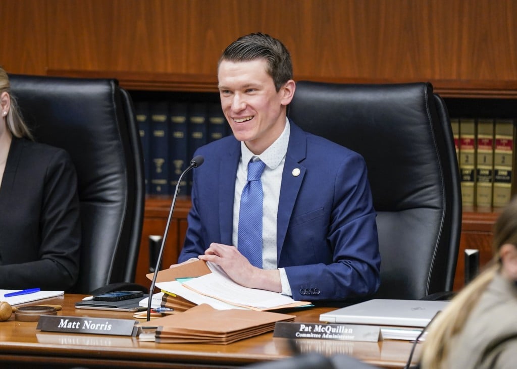 Minnesota State Representative Matt Norris smiles while seated at a committee desk with files and a microphone.