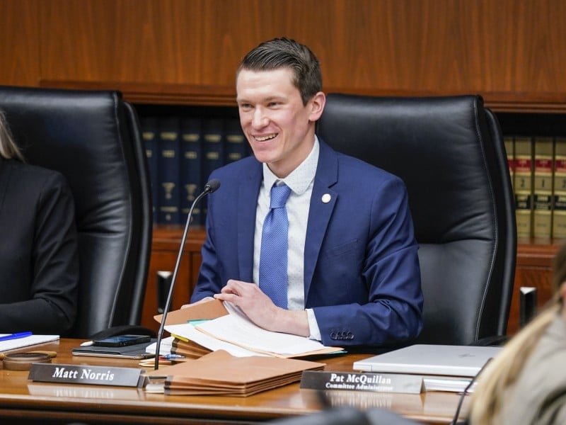 Minnesota State Representative Matt Norris smiles while seated at a committee desk with files and a microphone.